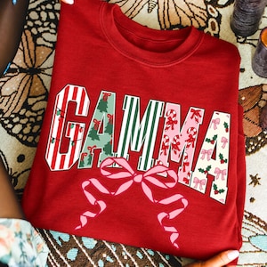Könnte beinhalten: Ein rotes Sweatshirt mit Rundhalsausschnitt und dem Wort GAMMA in großen Blockbuchstaben. Jeder Buchstabe hat ein einzigartiges Weihnachtsmuster, darunter rot-weiße Streifen mit Lichtern, grüne Tannenbäume, grüne Streifen, Zuckerstangen und rosa Schleifen.