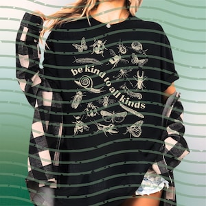 May include: Black t-shirt featuring white illustrations of insects such as butterflies, beetles, and a snail, with the text "be kind to all kinds". The shirt is worn with a black and white checked shirt. The design is a nature-themed graphic.