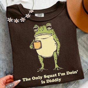 Könnte beinhalten: Ein dunkelbraunes T-Shirt mit einer Grafik eines grünen Frosches, der eine gelbe Tasse hält. Unter dem Frosch steht der Text: "The Only Squat I'm Doin' Is Diddly". Das T-Shirt hat einen Rundhalsausschnitt und ein "Comfort Colors"-Etikett.