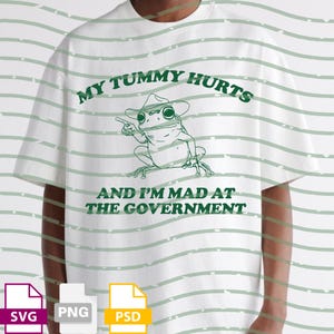 May include: White t-shirt with a green graphic of a frog wearing a cowboy hat, pointing. Text above reads "MY TUMMY HURTS" and below "AND I'M MAD AT THE GOVERNMENT". The shirt has a relaxed fit.