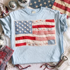 May include: Light blue t-shirt featuring a patchwork American flag design. The flag incorporates red and white stripes with floral, gingham, and polka dot patterns. White stars are sewn onto a blue background, creating a patriotic theme.
