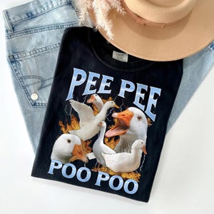 May include: Black graphic tee featuring the text "PEE PEE POO POO" in blue lettering with white geese illustrations. The shirt is laid on top of light wash denim jeans and a tan hat.