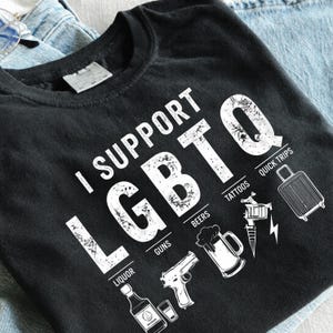 May include: A black t-shirt featuring white distressed text that reads I SUPPORT LGBTQ. Below the letters are icons representing LIQUOR, GUNS, BEERS, TATTOOS, and QUICK TRIPS, depicted as a bottle, a handgun, a beer mug, a tattoo machine, and a suitcase.