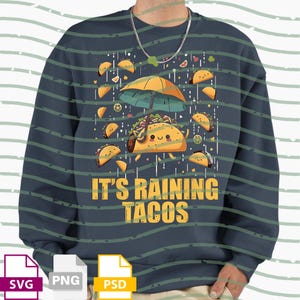 Puede incluir: Sudadera azul marino con un gráfico caprichoso de un taco bajo un paraguas, rodeado de tacos cayendo y el texto "IT'S RAINING TACOS" en amarillo. El diseño es lúdico y temático de comida.