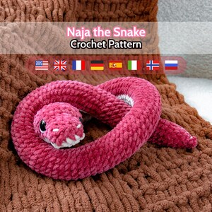 Puede incluir: Un juguete de serpiente rosa de felpa hecho a mano con ojos negros y boca blanca, enrollado sobre una superficie marrón texturizada. El texto "Naja the Snake Crochet Pattern" es visible.