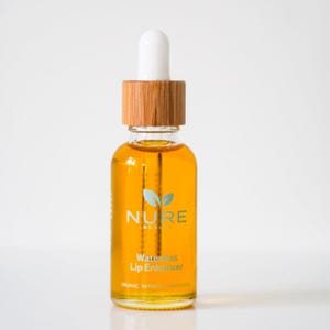 May include: A glass bottle of NURE BEAUTY Waterless Lip Enhancer. The bottle has a wooden cap and a white dropper. The amber-coloured liquid is visible inside. The label displays the brand name and product description in a modern font.