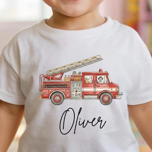 May include: A white t-shirt featuring a red and beige fire truck graphic with a ladder. The name "Oliver" is written in cursive below the truck. The child is smiling. The shirt is from Posh Pixie Little.