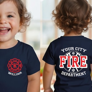 May include: Navy blue toddler t-shirt with a red and white fire department emblem on the front and back. The front of the shirt has the name "William" below the emblem. The back of the shirt says "Your City Fire Department".