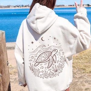 May include: Cream-coloured hoodie featuring a sea turtle graphic on the back. The design includes a turtle, waves, stars, and aquatic plants in brown. The hoodie has a hood and long sleeves.