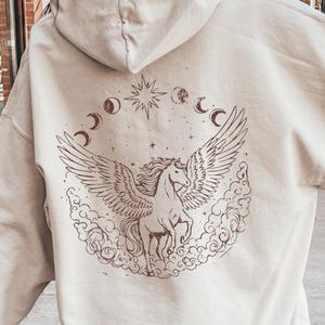 May include: Cream-colored hoodie with a drawstring hood. The back features a brown graphic of a winged horse, surrounded by clouds, moons, and stars. The hoodie is a relaxed fit, suitable for casual wear.