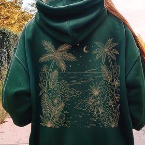 May include: A dark green hooded sweatshirt featuring a gold-colored line art graphic on the back. The design depicts a tropical beach scene with palm trees, monstera leaves, ferns, flowers, a crescent moon, and stars arranged in a square frame.