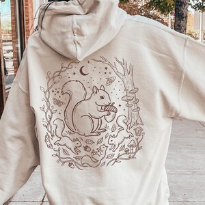 May include: Cream-colored hoodie with a brown illustration of a squirrel holding an acorn, surrounded by other squirrels, leaves, and mushrooms. The design includes a crescent moon and stars. The hoodie has a drawstring hood and a kangaroo pocket.
