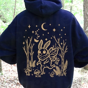 May include: A dark navy blue hooded sweatshirt featuring a gold-colored graphic on the back. The design depicts a moth-like creature with wings, surrounded by trees, mushrooms, a crescent moon, stars, and a small butterfly in a forest.