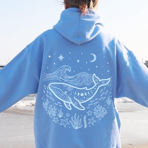 Op de afbeelding: Een lichtblauwe hoodie met een witte lijntekening op de achterkant. Het ontwerp toont een walvis omringd door oceaangolven, koraal, zeesterren en kleine vissen, met een maansikkel en sterren boven de walvis afgebeeld.