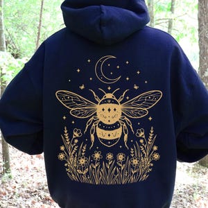 May include: A dark navy blue hooded sweatshirt featuring a gold-colored graphic on the back. The design displays a large bee centered above a field of wildflowers, with a crescent moon and small stars positioned at the top of the illustration.