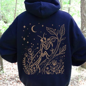 May include: Navy blue hoodie with a gold illustration of a praying mantis, moon, stars, and floral elements on the back. The design is centered and detailed, creating a nature-inspired aesthetic. The hoodie has a relaxed fit and a hood.