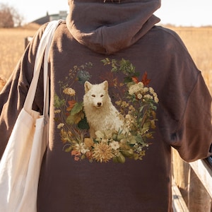 May include: A brown hoodie featuring a white wolf illustration surrounded by flowers and foliage. The wolf is the focal point, set against a backdrop of a natural landscape. The hoodie is a comfortable, casual garment.