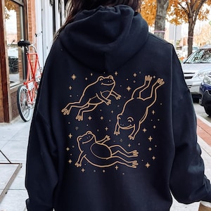 Può includere: Felpa con cappuccio nera con un'illustrazione dorata di tre rane e stelle sul retro. Le rane sono in diverse pose. La felpa ha un cappuccio e maniche lunghe.