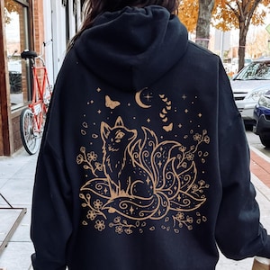 Peut inclure: Sweat à capuche noir avec une illustration dorée d'un renard à neuf queues, entouré de fleurs, de papillons et d'un croissant de lune. Le sweat à capuche a une capuche à cordon et des manches longues.