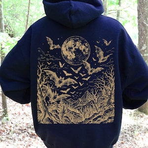 May include: A dark navy blue hooded sweatshirt featuring a large gold-colored graphic print on the back. The design depicts a full moon, numerous flying bats, bare trees, and a mountain landscape in a detailed, etched-style illustration.
