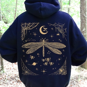May include: A dark navy blue hooded sweatshirt featuring a large gold-colored graphic on the back. The design displays a central dragonfly surrounded by small bees, a crescent moon, stars, planets, and spider webs in the corners of the rectangular print.