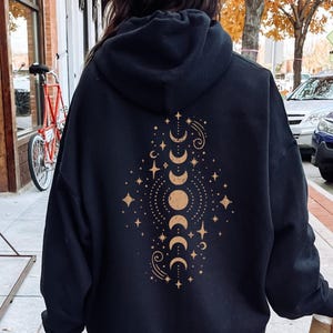 May include: Black hooded sweatshirt with a celestial design on the back. The design features phases of the moon, stars, and other celestial elements in a gold colour. The sweatshirt is a casual style.