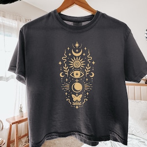 May include: A charcoal grey short-sleeve t-shirt featuring a vertical gold-coloured graphic print. The design includes celestial elements such as a sun, moon, stars, an eye, a butterfly, and botanical sprigs arranged in a symmetrical pattern.