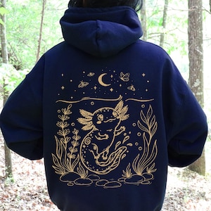 May include: A dark navy blue hooded sweatshirt featuring a gold-coloured line art graphic on the back. The design depicts an axolotl swimming among aquatic plants and rocks, with a crescent moon, stars, and small moths positioned above it.