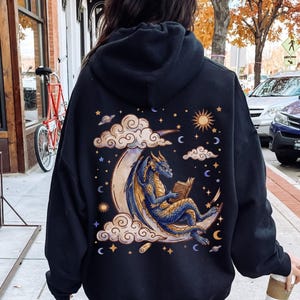 Può includere: Una felpa nera con cappuccio caratterizzata da una grande grafica sul retro. Il disegno raffigura un drago blu con ali dorate seduto su una luna crescente, intento a leggere un libro tra nuvole stilizzate, stelle, un sole e pianeti dorati e blu.