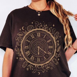 May include: Dark chocolate brown t-shirt with a clock design featuring woodland creatures and Roman numerals. The clock face is surrounded by a wreath of leaves, mushrooms, and animals. The text "Dark Chocolate" is at the bottom.