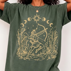 May include: A moss green short-sleeve t-shirt featuring a gold-colored graphic print of an archer aiming a bow and arrow within a forest setting, surrounded by a celestial moon phase cycle and a central star design on the front chest area.