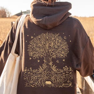 May include: A dark brown hooded sweatshirt featuring a gold-colored line art graphic on the back. The design depicts a large tree with fruits, flowers, birds, and insects, with visible roots extending underground to show a small burrowing animal.