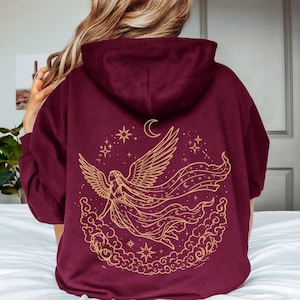 May include: A maroon hooded sweatshirt featuring a large gold-colored line art graphic on the back. The design depicts a winged figure with long flowing hair and robes, surrounded by clouds, stars, and a crescent moon in a circular arrangement.