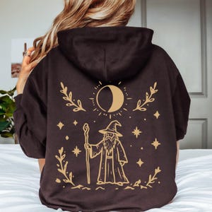 May include: A dark brown hooded sweatshirt featuring a light tan graphic print on the back. The design displays a wizard holding a staff, surrounded by a crescent moon, stars, and leafy branches. The hoodie has long sleeves and a hood.