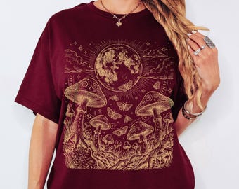 May include: A maroon short-sleeved t-shirt featuring a gold-toned graphic print of a large moon, several mushrooms, and various butterflies. The design includes intricate line work, celestial stars, and small cloud motifs across the front.