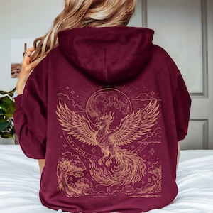 May include: Burgundy hoodie with a gold phoenix design on the back. The design features a phoenix rising from flames, a full moon, stars, and stylized trees. The hoodie has a hood and a relaxed fit.
