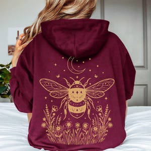 May include: A maroon hooded sweatshirt featuring a large gold-colored graphic on the back. The design displays a central bumblebee surrounded by a crescent moon, small stars, and a field of wildflowers at the bottom, all rendered in fine line art.