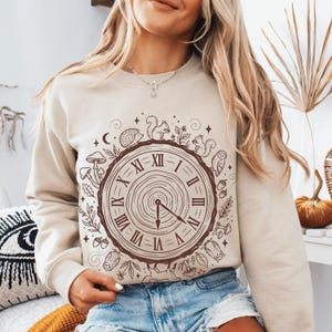 May include: Beige sweatshirt featuring a brown clock design with Roman numerals and woodland creatures like squirrels, owls, and hedgehogs. The clock hands point to approximately 5 o'clock. The design is surrounded by leaves, mushrooms, and acorns.