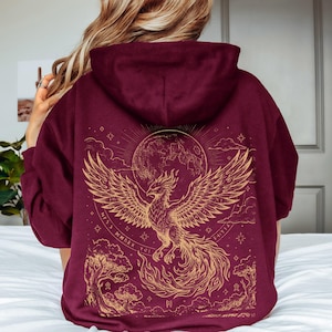 Op de afbeelding: Een bordeauxrode hoodie met een grote goudkleurige afbeelding op de achterkant. Het ontwerp toont een feniks met gespreide vleugels, een volle maan, wolken en bomen, omringd door cirkelvormige patronen en kleine stervormige symbolen.