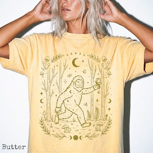 May include: Pale yellow t-shirt with a Bigfoot graphic holding a butterfly, set in a forest scene with trees, moons, and stars. The design is in shades of green. The word "Butter" is printed at the bottom of the shirt.