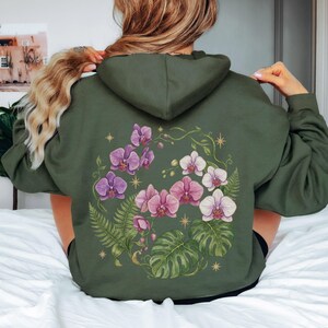 Puede incluir: Una sudadera con capucha de color verde oliva que presenta un gran gr&aacute;fico en la espalda de orqu&iacute;deas moradas y blancas, frondas de helecho verdes y hojas de monstera. El dise&ntilde;o incluye peque&ntilde;os s&iacute;mbolos de estrellas doradas en c&iacute;rculo.