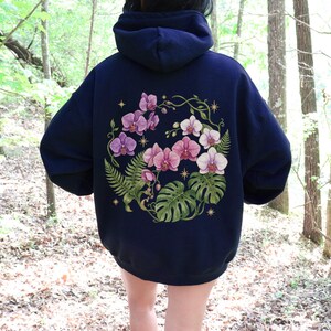 Puede incluir: Una sudadera con capucha azul marino oscuro que presenta un gran gr&aacute;fico en la espalda de orqu&iacute;deas rosas y blancas, hojas de monstera verdes y frondas de helecho dispuestas en un patr&oacute;n circular, con peque&ntilde;as estrellas doradas.
