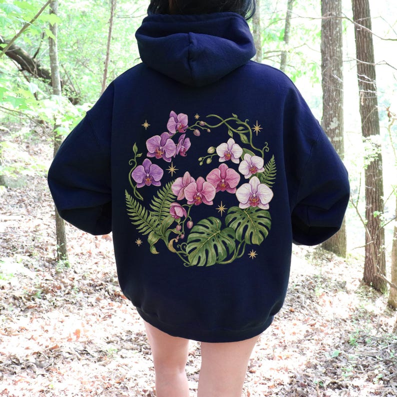 Puede incluir: Una sudadera con capucha azul marino oscuro que presenta un gran gr&aacute;fico en la espalda de orqu&iacute;deas rosas y blancas, hojas de monstera verdes y frondas de helecho dispuestas en un patr&oacute;n circular, con peque&ntilde;as estrellas doradas.