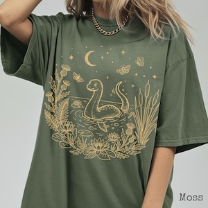 Pode incluir: T-shirt verde musgo com uma ilustração bege de uma cena lacustre. O desenho apresenta uma criatura semelhante ao monstro do lago Ness, uma lua, estrelas, borboletas e plantas aquáticas. Mangas curtas, corte folgado.