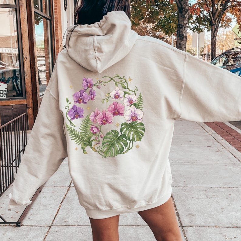 Puede incluir: Una sudadera con capucha de color beige que presenta un gran gr&aacute;fico circular en la espalda. El dise&ntilde;o muestra flores de orqu&iacute;dea moradas y blancas entrelazadas con hojas de helecho y monstera verdes, acentuadas por peque&ntilde;as estrellas amarillas.