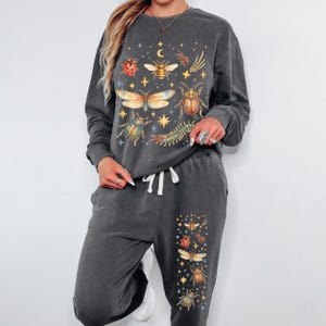 May include: A charcoal grey long-sleeve sweatshirt and matching sweatpants set featuring a colourful graphic print of various insects, including beetles, a bee, and a centipede, interspersed with yellow stars and a crescent moon on the chest and leg.