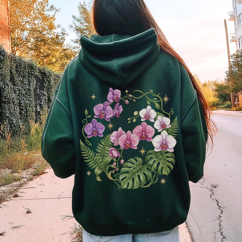 Puede incluir: Una sudadera con capucha de color verde oscuro que presenta un gran gr&aacute;fico en la espalda de orqu&iacute;deas moradas y blancas, frondas de helecho verdes y hojas de monstera dispuestas en un patr&oacute;n circular, con peque&ntilde;as estrellas amarillas de cuatro puntas.
