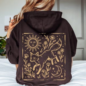 Könnte beinhalten: Ein dunkelbrauner Kapuzenpullover mit einer großen rechteckigen Grafik auf der Rückseite. Der Druck zeigt einen stilisierten Fuchs, umgeben von Blumen, einer Sonne mit Gesicht, einer Mondsichel und kleinen Sternen in hellbrauner Linienführung.