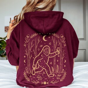 May include: A maroon hooded sweatshirt featuring a gold-colored graphic on the back. The design depicts a large creature walking through a forest with trees, mushrooms, butterflies, stars, and a crescent moon, all framed by a decorative border.