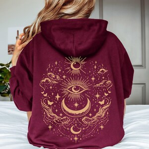May include: A deep burgundy hooded sweatshirt featuring a large, intricate gold-colored graphic on the back. The design includes a central eye, a crescent moon, stars, clouds, and butterflies arranged in a symmetrical, celestial-inspired pattern.